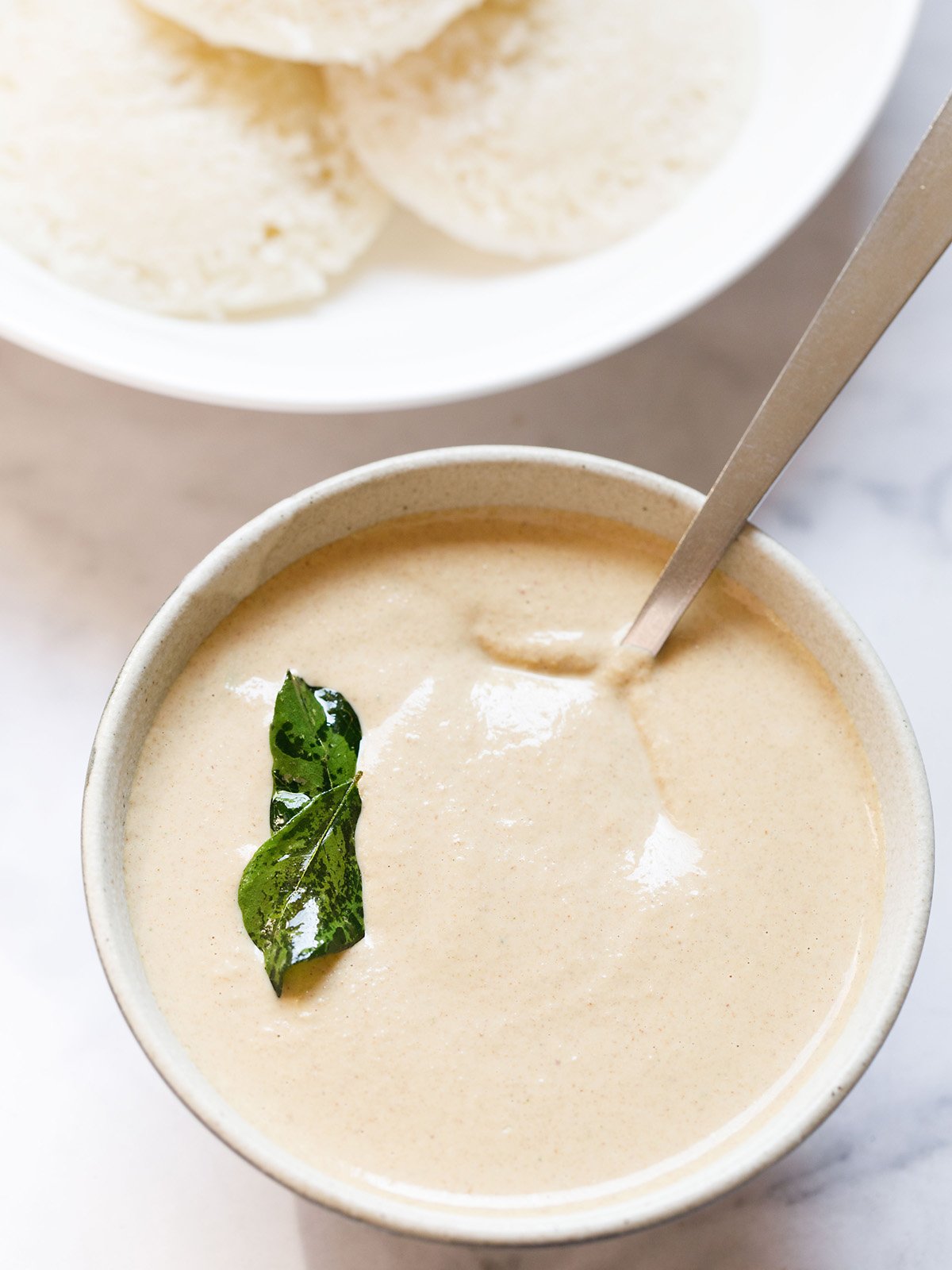 groundnut chutney in a ceramic bowl with fried curry leaves on it with a steel spoon on a white marble background