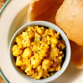 jeera aloo served in a ceramic bowl and puri on one side served in a cream enamel plate