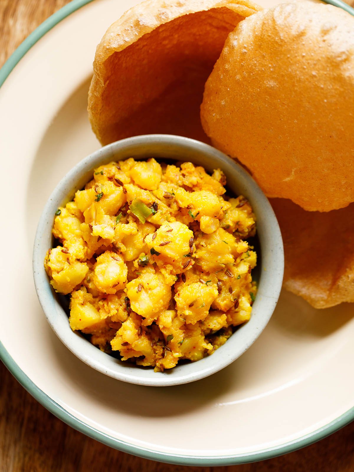 jeera aloo served in a ceramic bowl with a side of puri in a cream enamel plate.