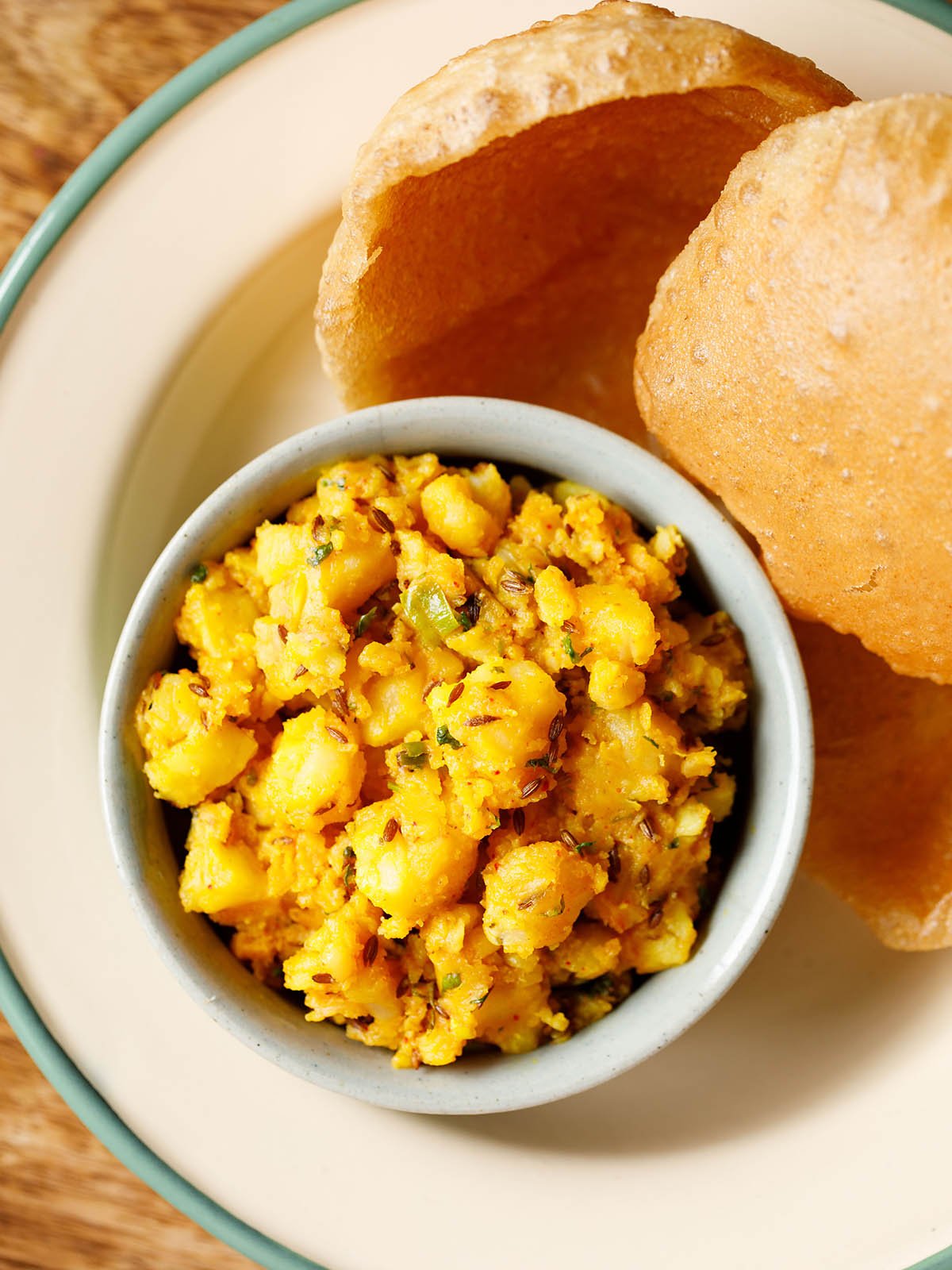 jeera aloo served in a ceramic bowl with a side of puri in a cream enamel plate