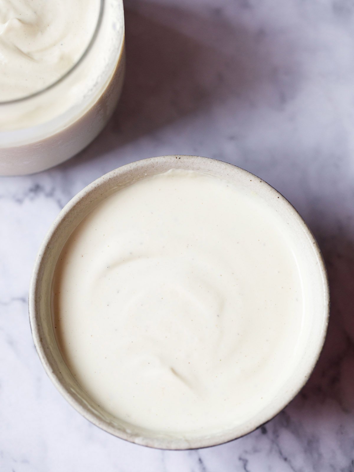 glass jar and small earthenware bowl with vegan mayonnaise on a grey and white marble tabletop