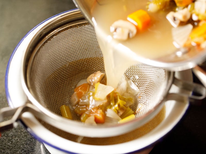 hand pouring cooked and cooled vegetable broth through a strainer