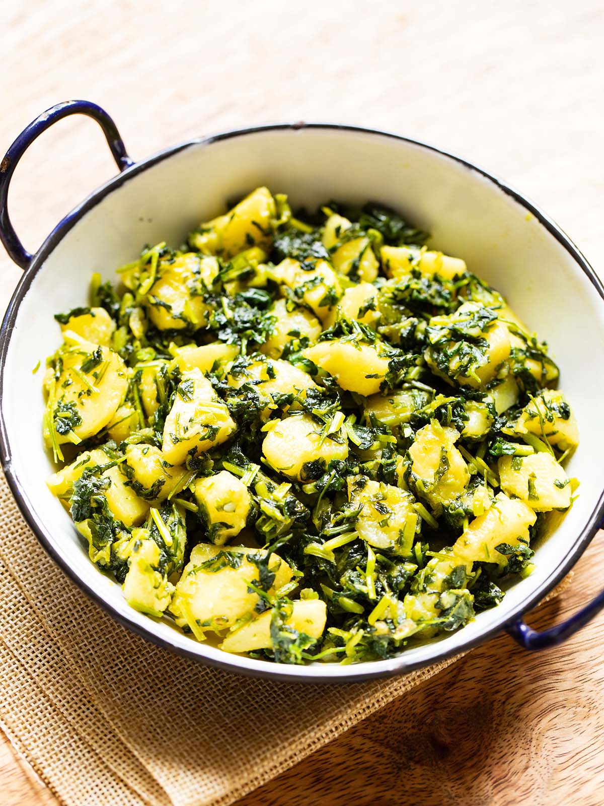 aloo methi in a enameled black rimmed small white wok on a brown jute napkin