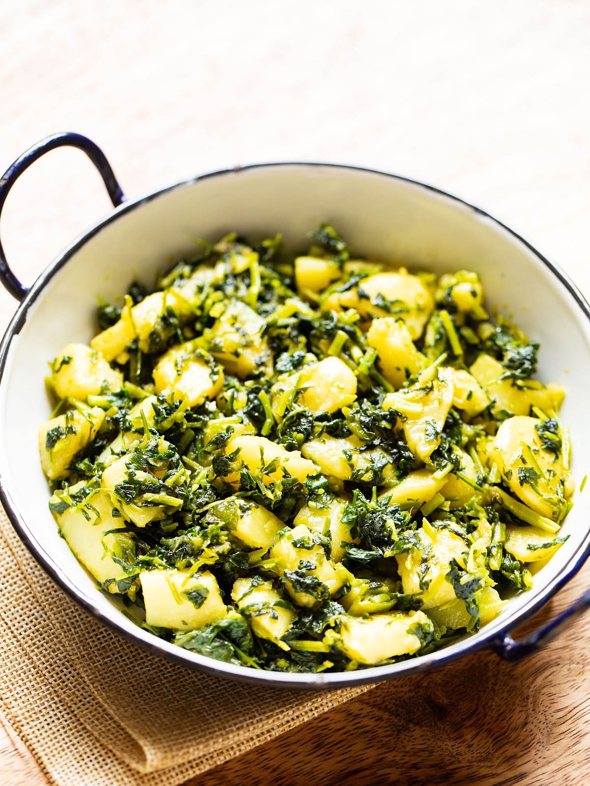 aloo methi in a enameled black rimmed small white wok on a brown jute napkin