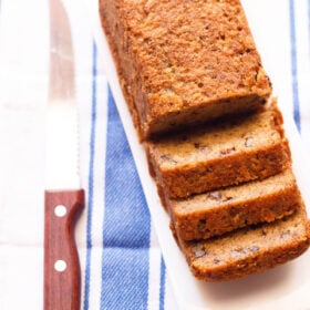 loaf of sliced eggless banana cake on white tray with serrated wooden knife on side