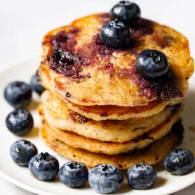 stack of blueberry pancakes with blueberries on top at sides on a white plate.
