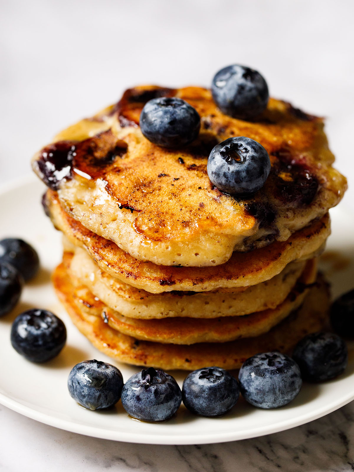 45 degree angle shot of 5 blueberry pancakes garnished with fresh blueberries and maple syrup on white plate