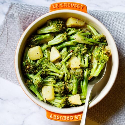 sauteed broccoli with potatoes in a le creuset cream ceramic pan with orange handles placed on a folded light gray jute fabric