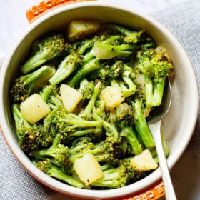 sauteed broccoli in a le creuset cream ceramic pan with orange handles with a spoon inside placed on a folded light gray jute fabric