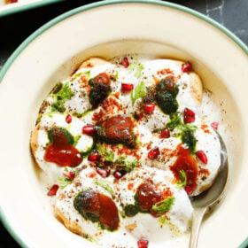 top shot of assembled dahi vada in a light green rimmed cream shallow plate topped with white curd, reddish tamarind chutney, green coriander chutney, pomegranate arils, chaat masala, cumin powder and coriander leaves