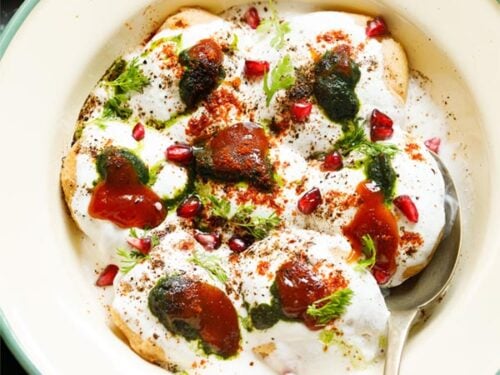 top shot of assembled dahi vada in a light green rimmed cream shallow plate topped with white curd, reddish tamarind chutney, green coriander chutney, pomegranate arils, chaat masala, cumin powder and coriander leaves