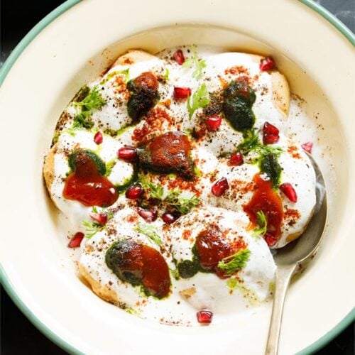 top shot of assembled dahi vada in a light green rimmed cream shallow plate topped with white curd, reddish tamarind chutney, green coriander chutney, pomegranate arils, chaat masala, cumin powder and coriander leaves