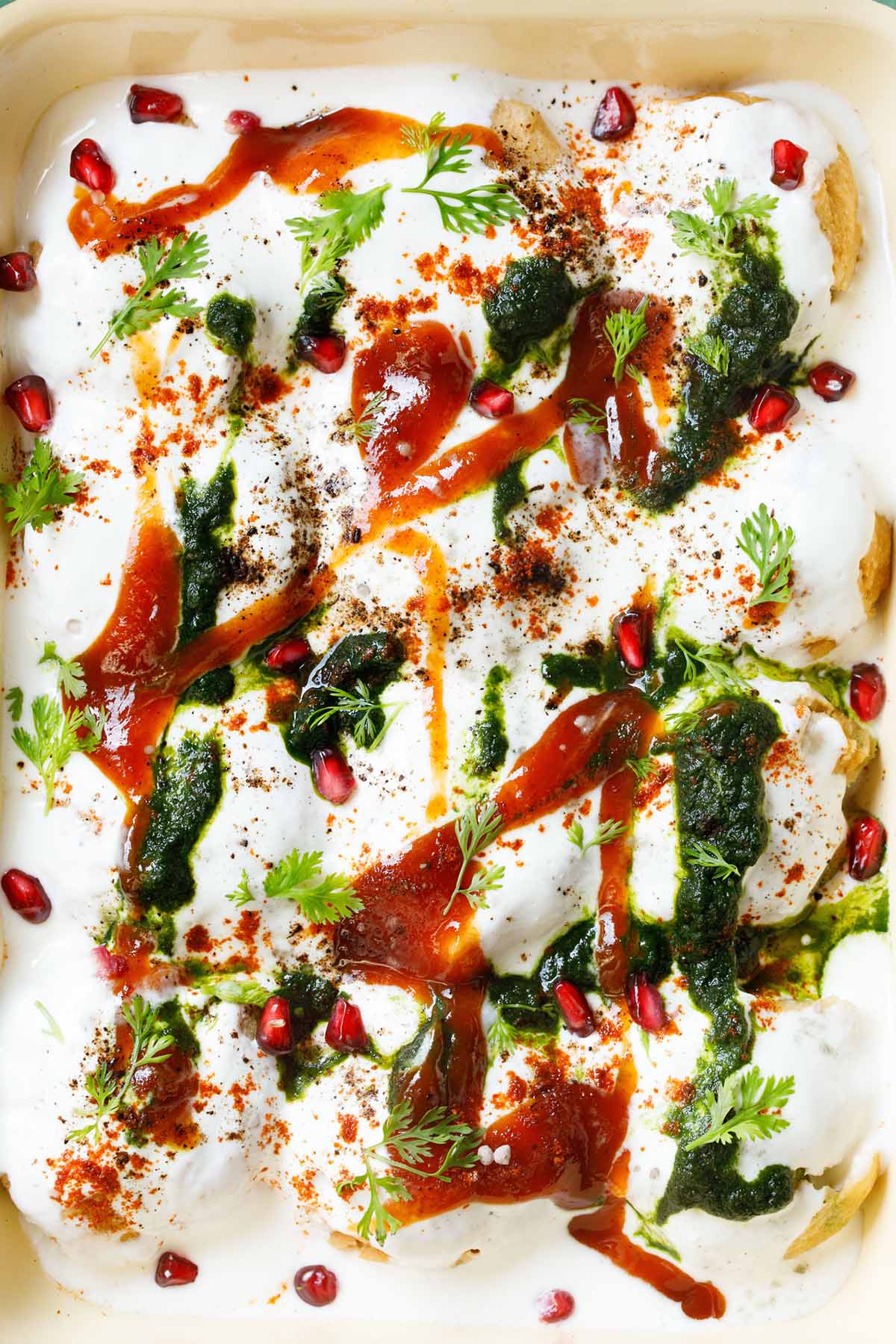 top closeup shot of assembled dahi vada in a cream tray topped with white curd, reddish tamarind chutney, green coriander chutney, pomegranate arils, chaat masala, cumin powder and coriander leaves
