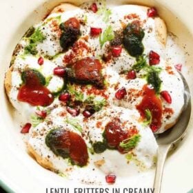 top shot of assembled dahi vada in a light green rimmed cream shallow plate topped with white curd, reddish tamarind chutney, green coriander chutney, pomegranate arils, chaat masala, cumin powder and coriander leaves