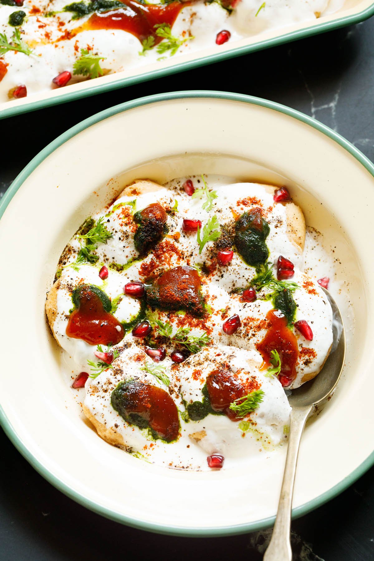 top shot of assembled dahi vada in a light green rimmed cream shallow plate topped with white curd, reddish tamarind chutney, green coriander chutney, pomegranate arils, chaat masala, cumin powder and coriander leaves