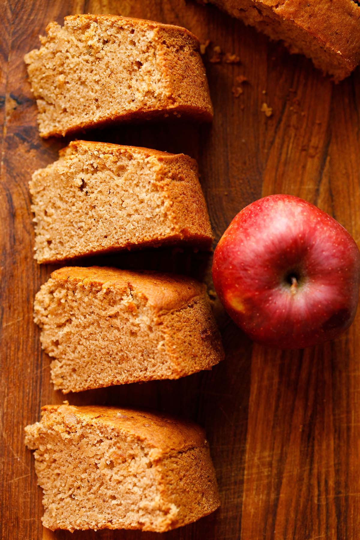 three slices of eggless apple cake on wooden table next to red apple