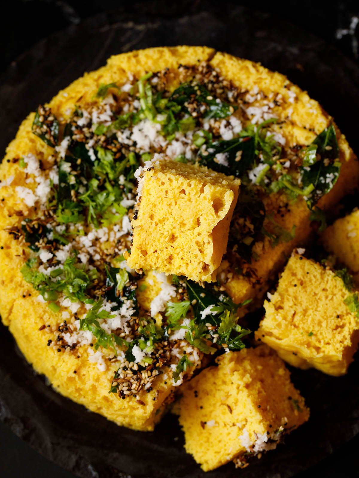 a tender soft block of khaman kept on top the round steamed cake with some khaman cubes at the side on a black granite tray