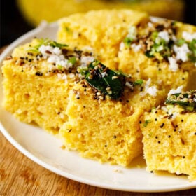 45 degree angle shot of khaman dhokla squares on a white plate garnished with cilantro, curry leaves and the fried curry leaves and spices mixture