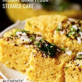 45 degree angle shot of khaman dhokla squares on a white plate garnished with cilantro, curry leaves and the fried curry leaves and spices mixture