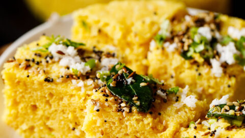 45 degree angle shot of khaman dhokla squares on a white plate garnished with cilantro, curry leaves and the fried curry leaves and spices mixture