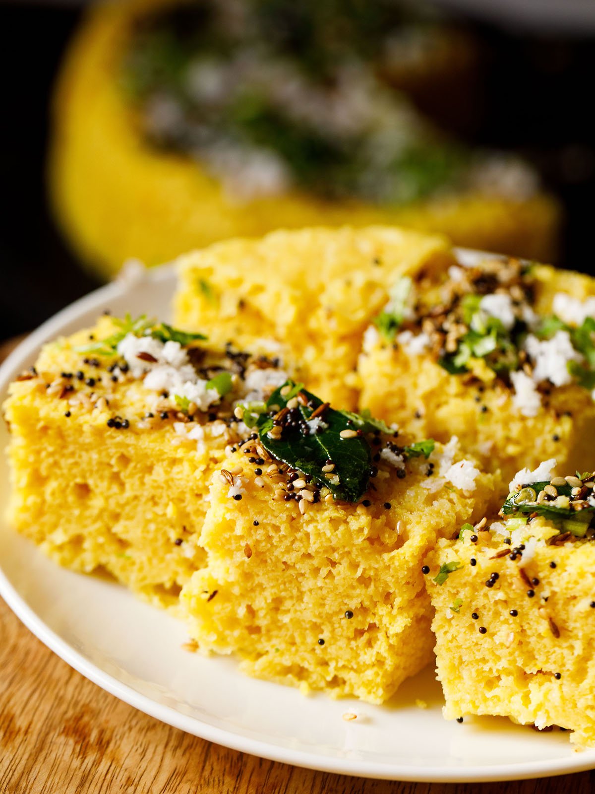 45 degree angle shot of khaman dhokla squares on a white plate garnished with cilantro, curry leaves and the fried curry leaves and spices mixture