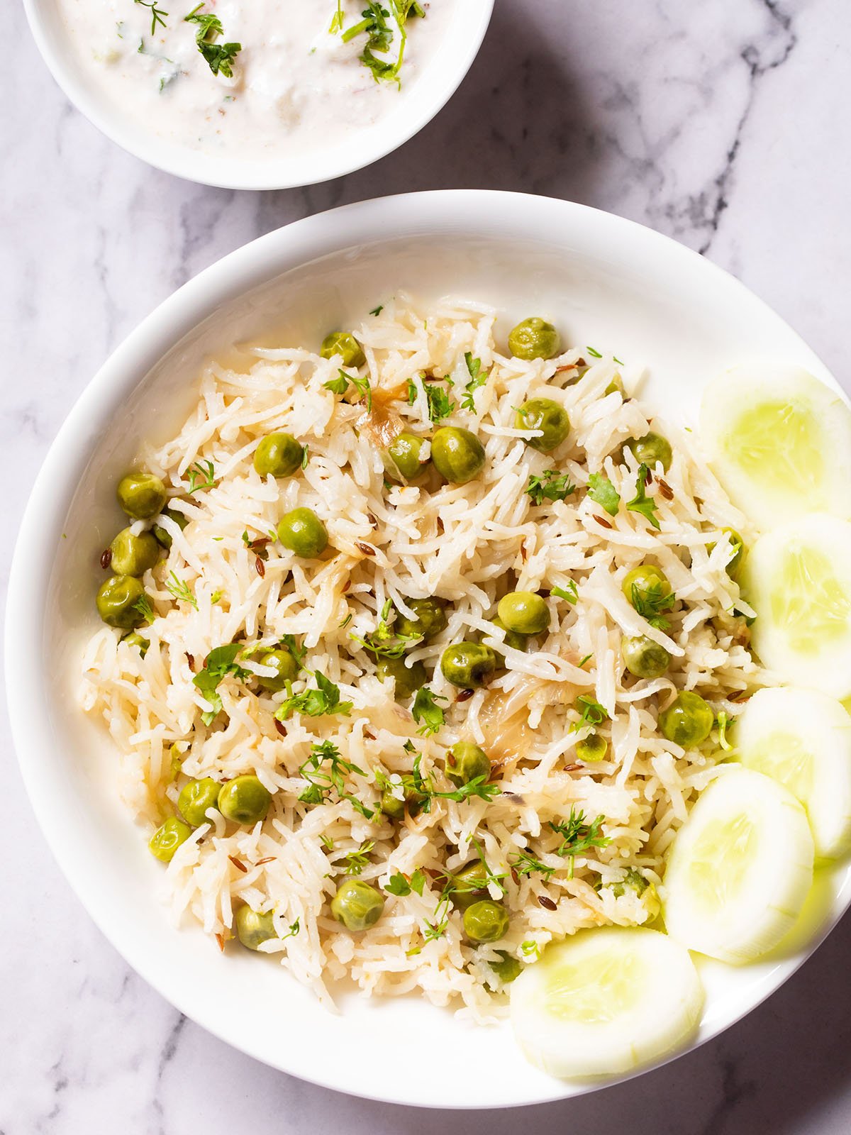 overhead shot of green peas pulao in a white shallow bowl with cucumber slices placed at the right side on a white marble table with a raita in white bowl at the top