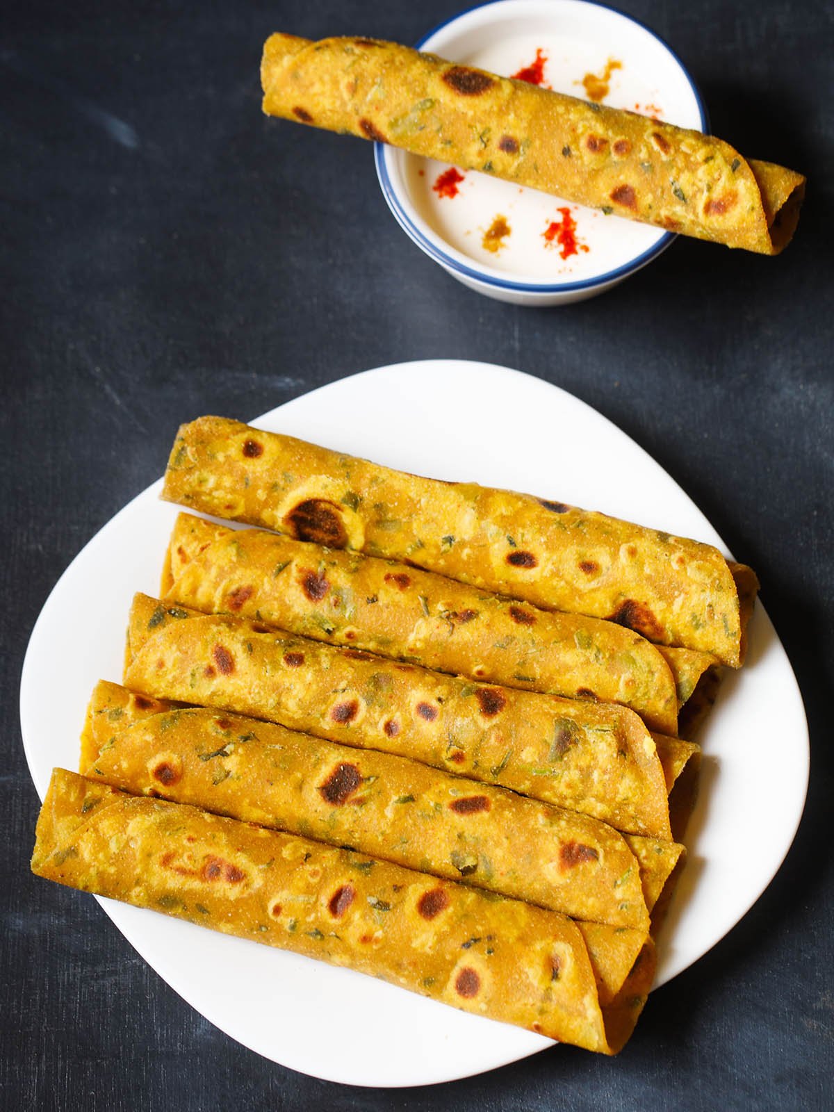 methi thepla rolls placed stacked vertically in white square plate on a dark blue black board