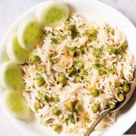 overhead shot of green peas pulao in a white shallow bowl with a brass spoon and cucumber slices placed at the left side on a white marble table