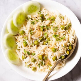 overhead shot of green peas pulao in a white shallow bowl with a brass spoon and cucumber slices placed at the left side on a white marble table