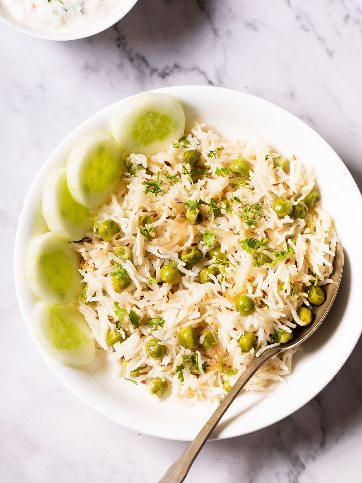 overhead shot of green peas pulao in a white shallow bowl with a brass spoon and cucumber slices placed at the left side on a white marble table