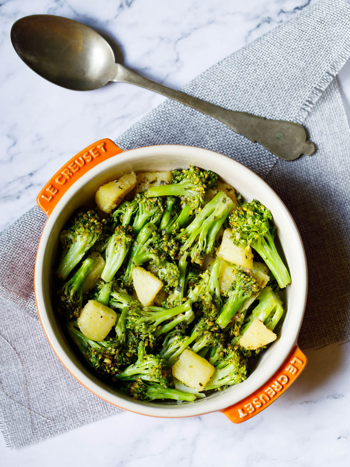 top view of sauteed broccoli in a le creuset cream ceramic pan with orange handles placed on a folded light gray jute fabric with an antique brass spoon on top