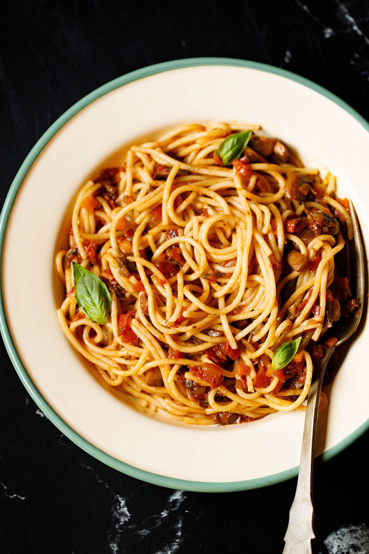 close up shot of spaghetti bolognese garnished with basil leaves in a green rimmed cream colored enameled plate with brass fork inside