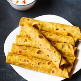 a methi thepla roll placed in a slant direction over five stacked methi thepla rolls on white plate on blue black board with a side of raita on top left