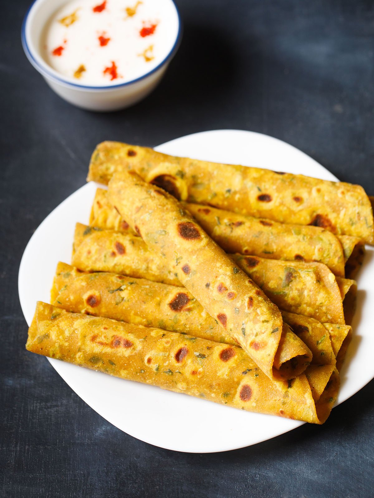 a methi thepla roll placed in a slant direction over five stacked methi thepla rolls on white plate on blue black board with a side of raita on top left