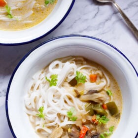 overhead shot of vegan tom kha (thai coconut soup) over rice noodles in a white bowl with a blue rim on a white marble table with a silver spoon