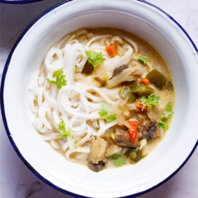 overhead shot of vegan tom kha (thai coconut soup) over rice noodles in a white bowl with a blue rim on a white marble table