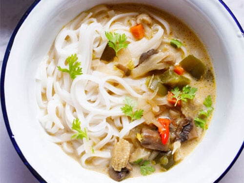 overhead shot of vegan tom kha (thai coconut soup) over rice noodles in a white bowl with a blue rim on a white marble table