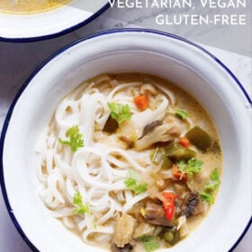 overhead shot of vegan tom kha (thai coconut soup) over rice noodles in a white bowl with a blue rim on a white marble table with a silver spoon with text layovers