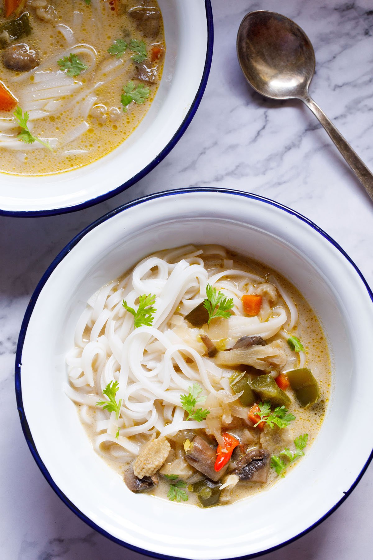 overhead shot of vegan tom kha (thai coconut soup) over rice noodles in a white bowl with a blue rim on a white marble table with a silver spoon