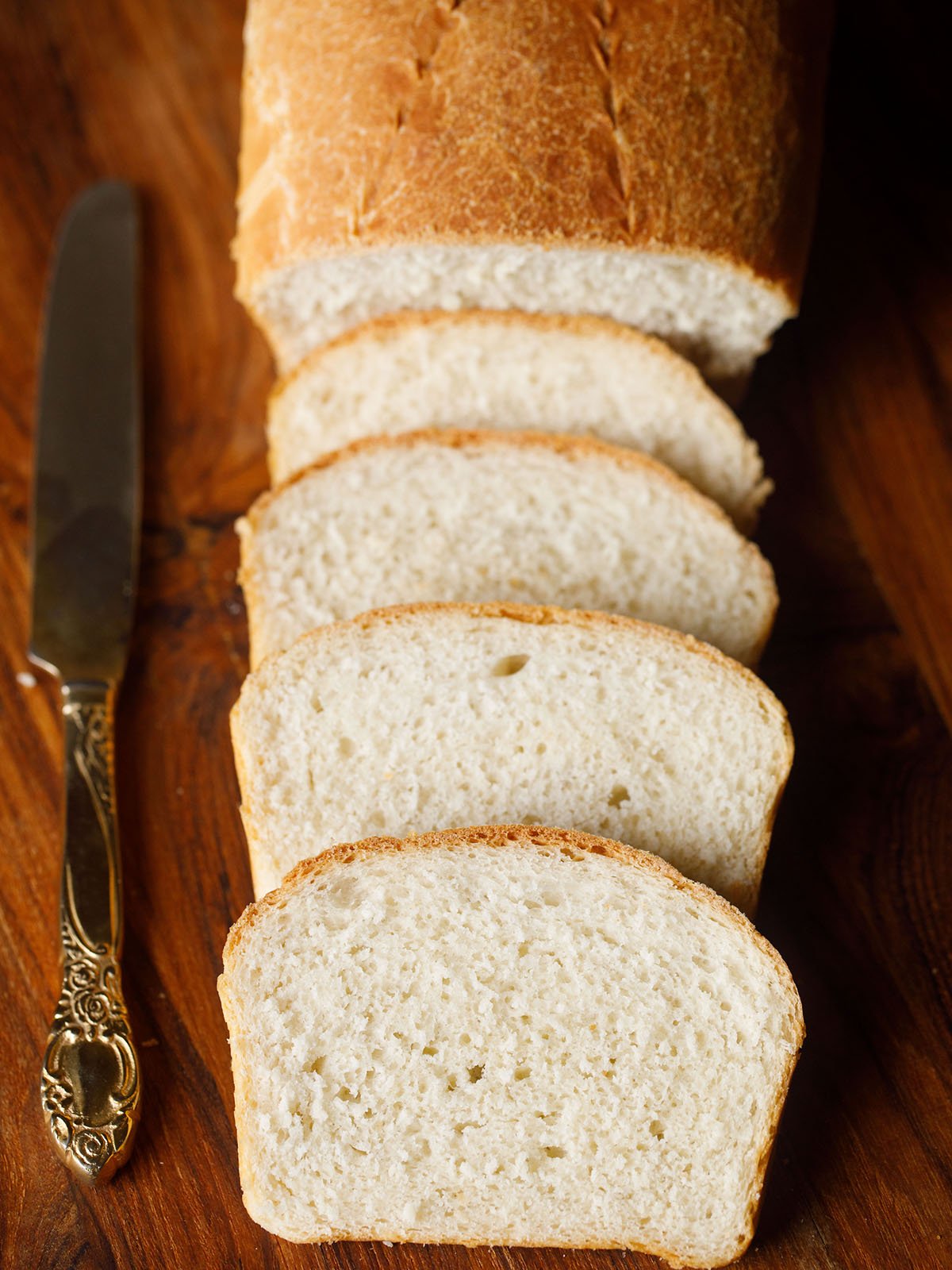 overheat shot of four slices of white bread with a partial view of the loaf with an brass butter knife on a brown wooden chopping board