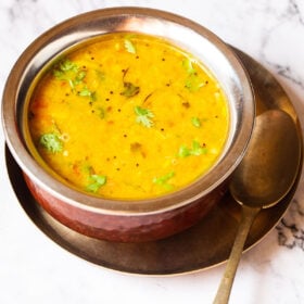 Dal fry in a copper pan with a brass spoon inside placed on a small brass plate on a white marble board