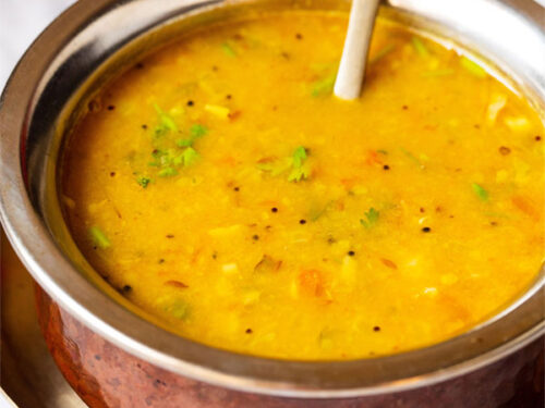 Close up photo of dal fry in a copper pan with a brass spoon inside placed on a small brass plate on a white marble board