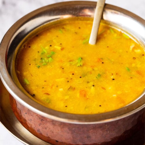 Close up photo of dal fry in a copper pan with a brass spoon inside placed on a small brass plate on a white marble board