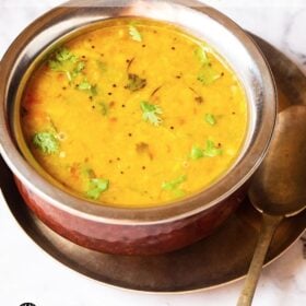 Dal fry in a copper pan with a brass spoon inside placed on a small brass plate on a white marble board