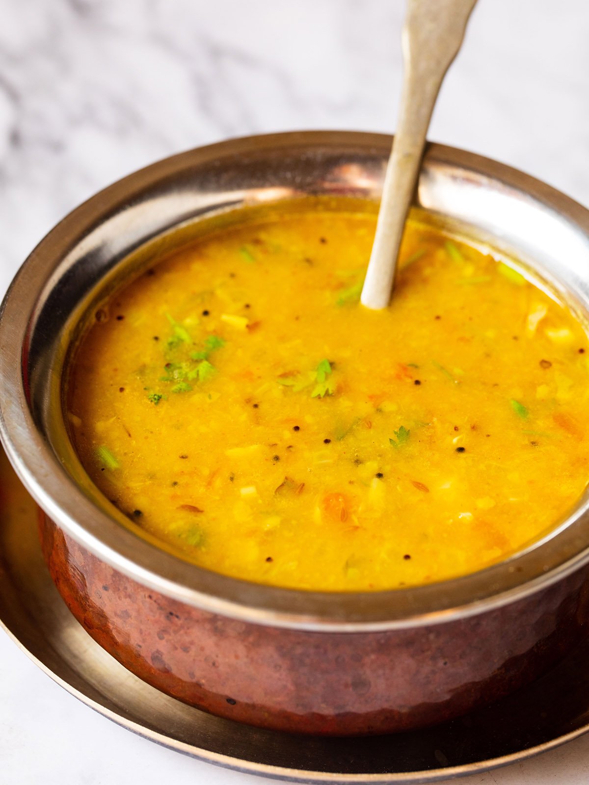 Close up photo of dal fry in a copper pan with a brass spoon inside placed on a small brass plate on a white marble board