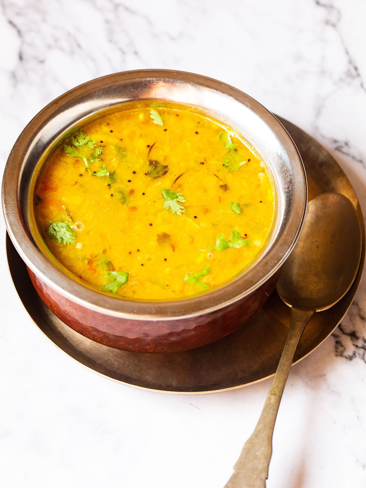 Dal fry in a copper pan with a brass spoon inside placed on a small brass plate on a white marble board