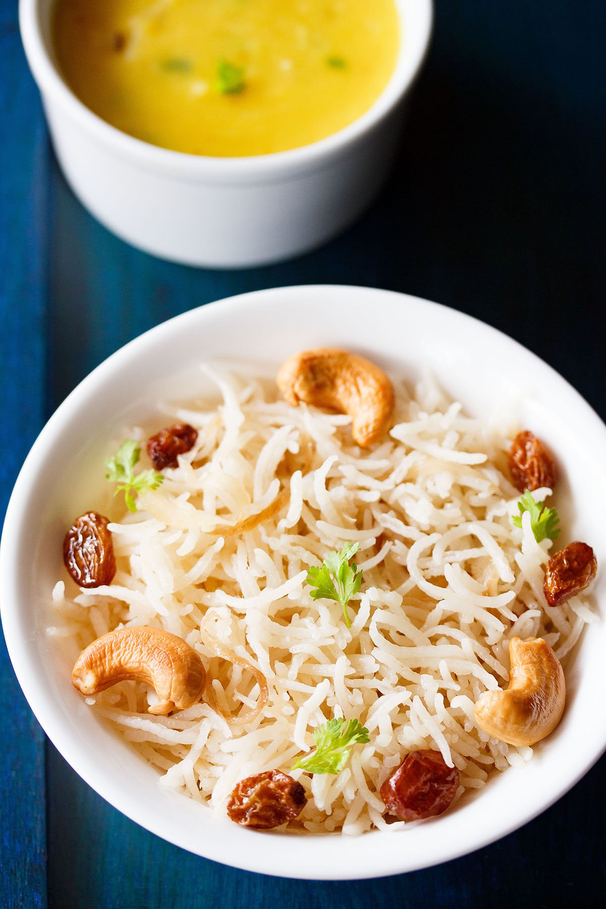 ghee rice topped with fried cashews, raisins, coriander leaves in a white bowl on a dark blue board