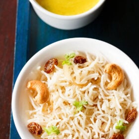 ghee rice topped with fried cashews, raisins, coriander leaves in a white bowl on a dark blue board with a bowl of yellow dal on top