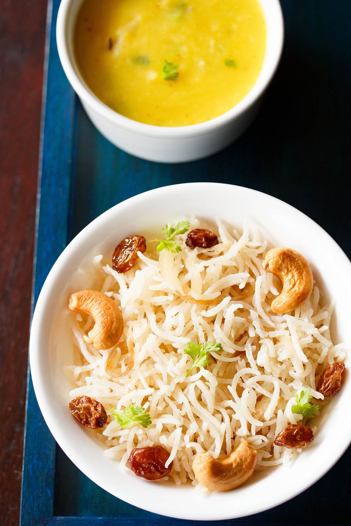 ghee rice topped with fried cashews, raisins, coriander leaves in a white bowl on a dark blue board with a bowl of yellow dal on top