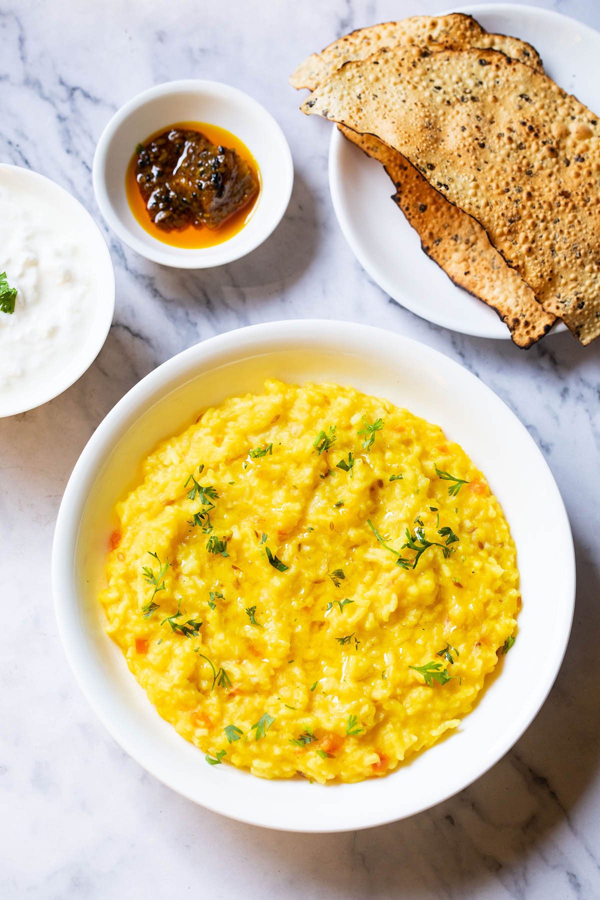 top shot of khichdi served in white bowl with sides of curd, mango pickle in white bowls and roasted papad served in a white plate